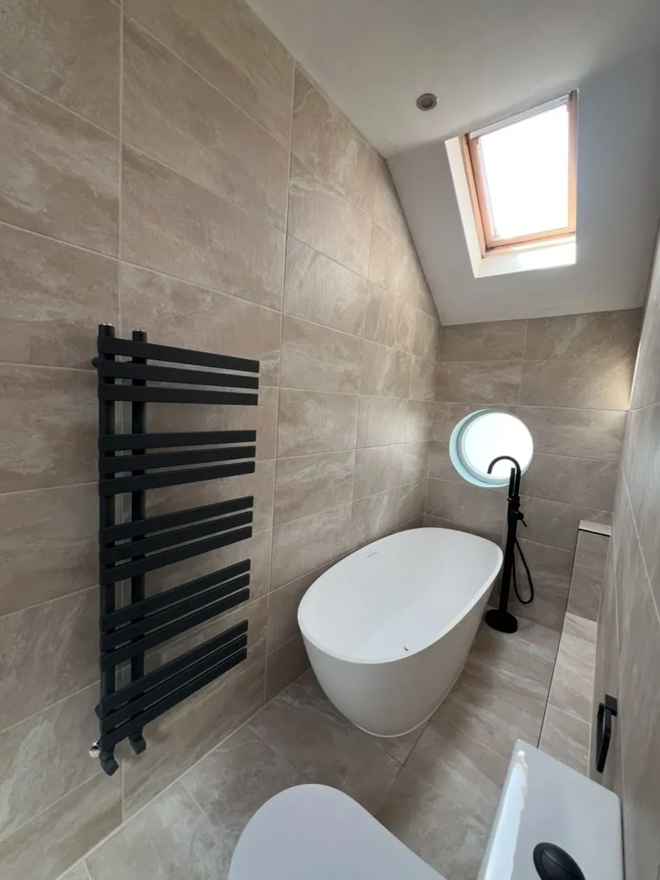 Loft bathroom installation in Reading with white freestanding bath, black towel radiator and stone-effect tiles